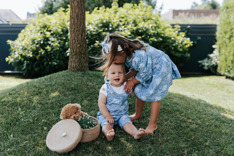 Deklica v pastelno modri obleki z drobnim cvetličnim potiskom in bratec v ujemajočem pajacku na travi.